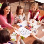 professora sorrindo em uma sala de aula com quatro crianças sorrindo, todas elas concentradas em um livro desenhado que a professora está mostrando. A sala de aula pode ter carteiras, quadro-negro, janelas e decorações temáticas de educação infantil. A professora pode estar apontando para alguma parte do livro enquanto as crianças estão olhando atentamente para as ilustrações. O clima é alegre e descontraído, com uma sensação de aprendizado divertido e envolvente.
